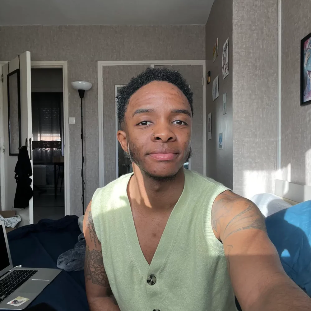 un selfie de Jay, un homme transgenre noir posant dans sa chambre à la lumière du soleil. Il porte un gilet vert pâle, a les cheveux noirs et court et sourie légèrement à la caméra.